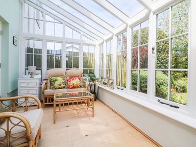A sunroom with rattan furniture and glass coffee table next to windows overlooking a garden at Home Farm Cottage in Fremington Devon