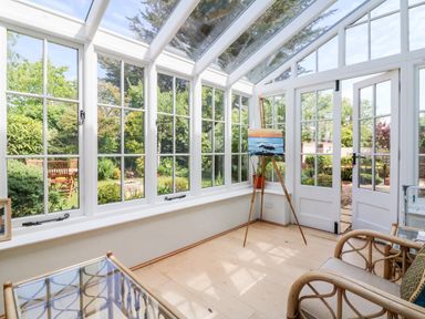 A sunroom with glass windows and doors a wooden easel with a painting and wicker chairs at Home Farm Cottage in Fremington Devon