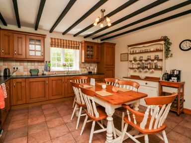 A kitchen with wooden cabinets a dining table with six chairs and shelves with cups at Home Farm Cottage in Fremington Devon