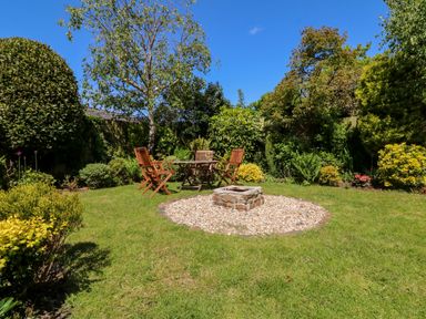A garden with a wooden table and chairs around a stone fire pit surrounded by plants and trees at Home Farm Cottage in Fremington Devon