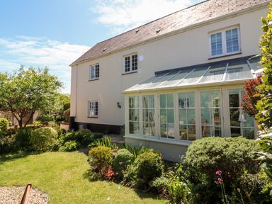 A garden with shrubs and trees beside a house with a glass conservatory at Home Farm Cottage in Fremington Devon