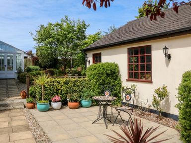 A patio area with a small table and two chairs next to potted plants and a cream building with red window frames at Home Farm Cottage in Fremington Devon