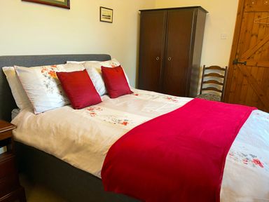 A bedroom with a bed that has floral bedding and red cushions with a wardrobe, wooden door, and chair at Home Farm Cottage in Fremington Devon