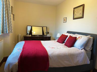 A bedroom with a bed with red pillows and a red runner a dresser with a three panel mirror and framed pictures on the wall at Home Farm Cottage in Fremington Devon