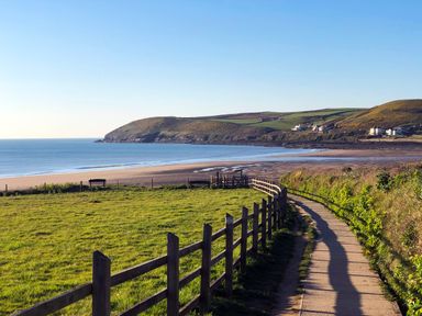 A coastal pathway with a wooden fence beside grassy fields leading to a sandy beach and hills in the background at Home Farm Cottage in Fremington Devon