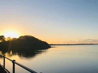 A sunset over a calm body of water with trees on the shore and a railing in the foreground at Home Farm Cottage in Fremington Devon