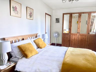 A bedroom with a wooden bed with white bedding and yellow pillows and blanket two bedside tables with lamps wooden wardrobe with mirrors and a standing fan at Home Farm Cottage in Fremington Devon