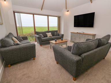 A living room with three gray sofas a coffee table a large window a wall-mounted television and a sideboard at Pol View in Gunwalloe near Helston