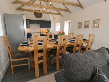 A dining area with a wooden table set for eight with plates glasses and tulips visible kitchen and clock on wall at Pol View Gunwalloe near Helston