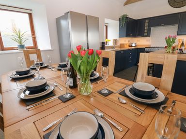 A kitchen and dining area with wooden chairs and a table set with bowls plates cutlery and glasses a vase of red tulips in the center a silver fridge and dark cabinets in the background at Pol View Gunwalloe near Helston