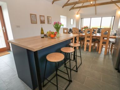 A kitchen island with three stools a bottle of wine a bowl of fruit and flowers with a dining table and chairs in the background at Pol View in Gunwalloe near Helston