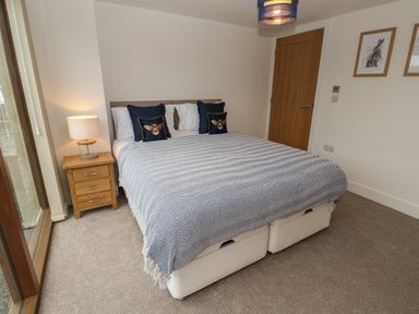 A bedroom with a double bed covered with a striped blanket two navy blue pillows with bee designs a wooden nightstand with a lamp a wooden door and two framed pictures of rabbits on the wall at Pol View in Gunwalloe near Helston