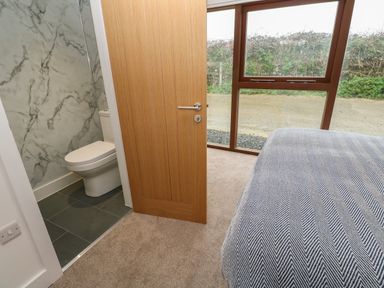 A bedroom showing a bed with a patterned cover near a wooden door leading to a bathroom with a toilet and marble wall and large windows looking outside at Pol View in Gunwalloe near Helston