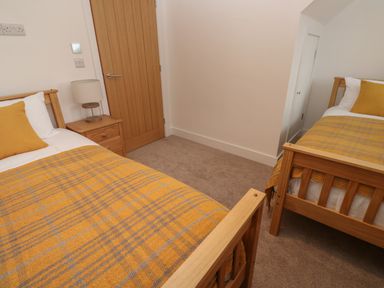 A bedroom with two single wooden beds with yellow and gray blankets and pillows a wooden door a nightstand with a lamp and white walls at Pol View Gunwalloe near Helston
