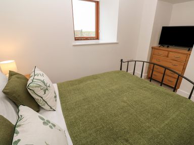 A bedroom with a bed with green bedding pillows a bedside lamp a wooden chest of drawers and a television at Pol View in Gunwalloe near Helston