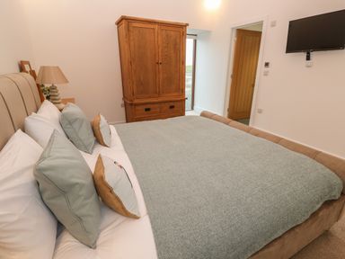 A bedroom with a bed with multiple pillows a wooden wardrobe a lamp a wall mounted tv and a door at Pol View Gunwalloe near Helston