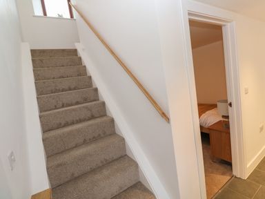 A carpeted staircase with a wooden handrail next to a doorway leading to a bedroom with a nightstand and lamp at Pol View Gunwalloe near Helston
