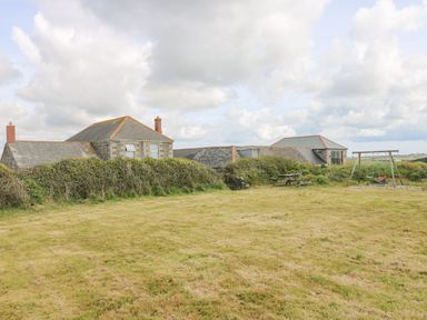 A large grassy yard with a picnic table and a swing set behind bushes near stone buildings at Pol View in Gunwalloe near Helston