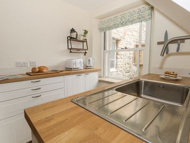 A kitchen with a stainless steel sink wooden countertops bread toaster kettle bread on a cutting board and a window at Bobbin Cottage in Swanage