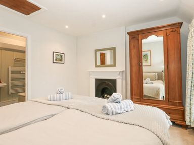 A bedroom with two beds with rolled towels a wooden wardrobe with a mirror a fireplace and a doorway to a bathroom at Bobbin Cottage in Swanage