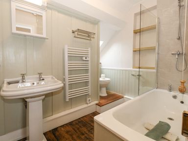 A bathroom with a pedestal sink a toilet on a raised platform a bathtub with a shower and wooden shelves at Bobbin Cottage in Swanage