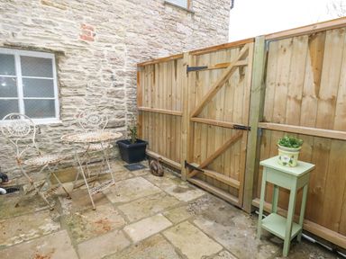 An outdoor patio area with a metal table and two chairs a wooden fence with a gate a stone wall window a small green table with a plant and a potted plant on the ground at Bobbin Cottage in Swanage