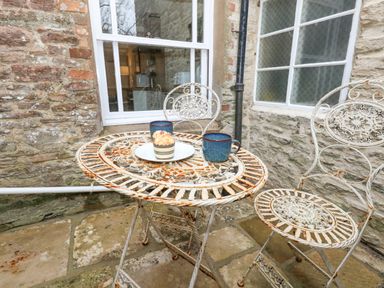 An outdoor stone patio with a round metal table and two metal chairs there are two blue mugs and a plate with a muffin on the table at Bobbin Cottage in Swanage