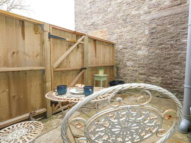 An outdoor patio with a metal table and chairs two cups and cupcakes on the table a wooden fence and stone wall in the background at Bobbin Cottage in Swanage