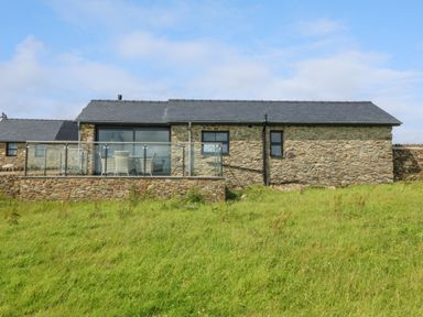 A stone house with a glass railing and outdoor furniture on a grass lawn at Tyddyn Bach in Rhydwyn