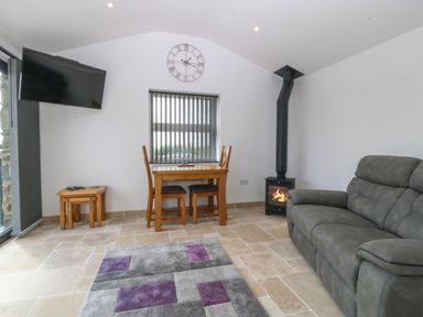 A living room with a grey sofa a small wooden dining table with two chairs a wall clock and a wood burning stove at Tyddyn Bach in Rhydwyn