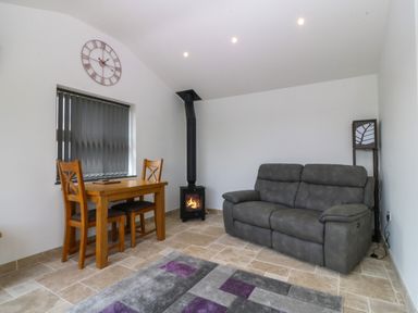 A small living room with a gray sofa a wooden table with two chairs a rug a black stove and a wall clock at Tyddyn Bach in Rhydwyn