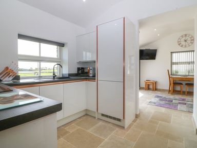 A kitchen area with white cabinets black countertop and a window next to a dining area with a wooden table chairs a wall clock and a television at Tyddyn Bach in Rhydwyn