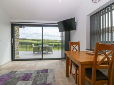 A dining area with a wooden table and chairs next to a window with vertical blinds and a wall-mounted TV near sliding glass doors leading to a patio with outdoor furniture and a view of green fields at Tyddyn Bach in Rhydwyn