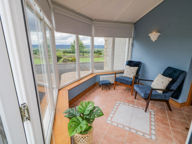 A corner room with two blue armchairs a small footstool a plant and large windows showing a garden view at Braeside Farm House in Cloughmills
