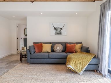 A living room with a grey sofa with orange and yellow cushions and a framed picture of a highland cow on the wall at 1 Caddaford Barns in Buckfastleigh
