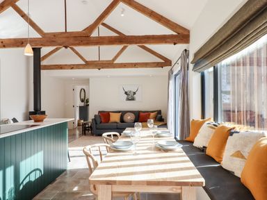 A dining area with a wooden table set with plates and glasses next to a bench with cushions and a living room with a sofa and wooden beams on the ceiling at 1 Caddaford Barns in Buckfastleigh
