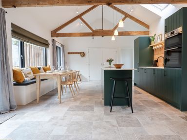 A kitchen and dining area with wooden beams overhead a dining table with chairs and a bench beside large windows at 1 Caddaford Barns in Buckfastleigh