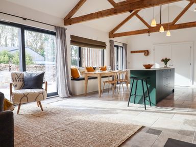 An open kitchen and dining area with wooden ceiling beams large windows a rug and a kitchen island at 1 Caddaford Barns in Buckfastleigh