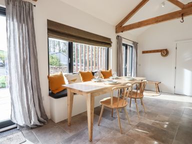 A dining area with a wooden table set with plates and glasses wooden chairs bench seating with cushions large windows with curtains and exposed wooden beams in the ceiling at 1 Caddaford Barns Buckfastleigh