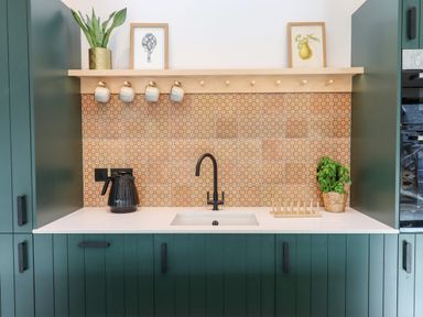 A kitchen sink area with green cabinets a black faucet a black kettle mugs on a wooden rack and framed pictures at 1 Caddaford Barns in Buckfastleigh