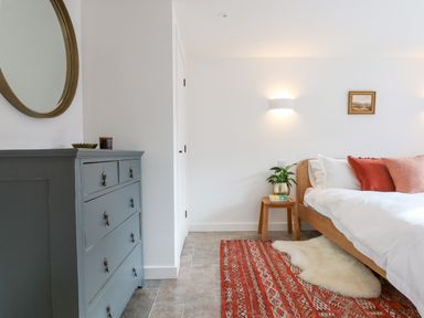 A bedroom with a wooden bed and white bedding a wooden nightstand with a plant a red patterned rug a gray dresser with a round mirror on the wall at 1 Caddaford Barns in Buckfastleigh