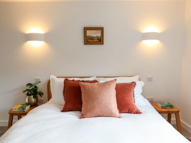 A bedroom with a bed with pillows two wall lights a picture and bedside tables with books and a plant at 1 Caddaford Barns in Buckfastleigh