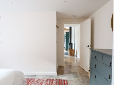 A room with a white bed corner a blue chest of drawers a red patterned rug and an open door leading to a hallway with a wood stove and basket at 1 Caddaford Barns in Buckfastleigh