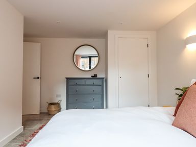 A bedroom with a bed white bedding a gray chest of drawers a round mirror and two closed doors at 1 Caddaford Barns in Buckfastleigh