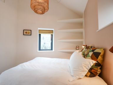 A bedroom with a bed with patterned pillows a window with a blind and corner shelves at 1 Caddaford Barns in Buckfastleigh