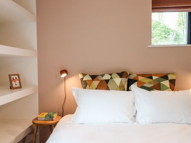 A bedroom with a bed with white pillows and patterned cushions beside a small wooden side table with books at 1 Caddaford Barns Buckfastleigh