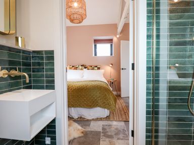 A bedroom with a bed and bedside table seen through a doorway from a bathroom with a sink and green tiled walls at 1 Caddaford Barns in Buckfastleigh