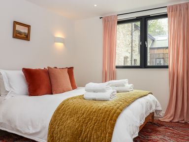 A bedroom with a bed with white linens orange pillows and a mustard blanket under a window with pink curtains at 1 Caddaford Barns Buckfastleigh