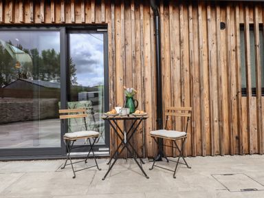 A small outdoor table with two chairs on a stone patio against a wooden wall at 1 Caddaford Barns in Buckfastleigh