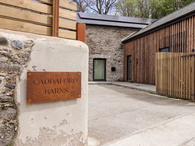A driveway with a stone wall and a metal sign reading caddaford barns next to modern barn buildings at 1 Caddaford Barns Buckfastleigh